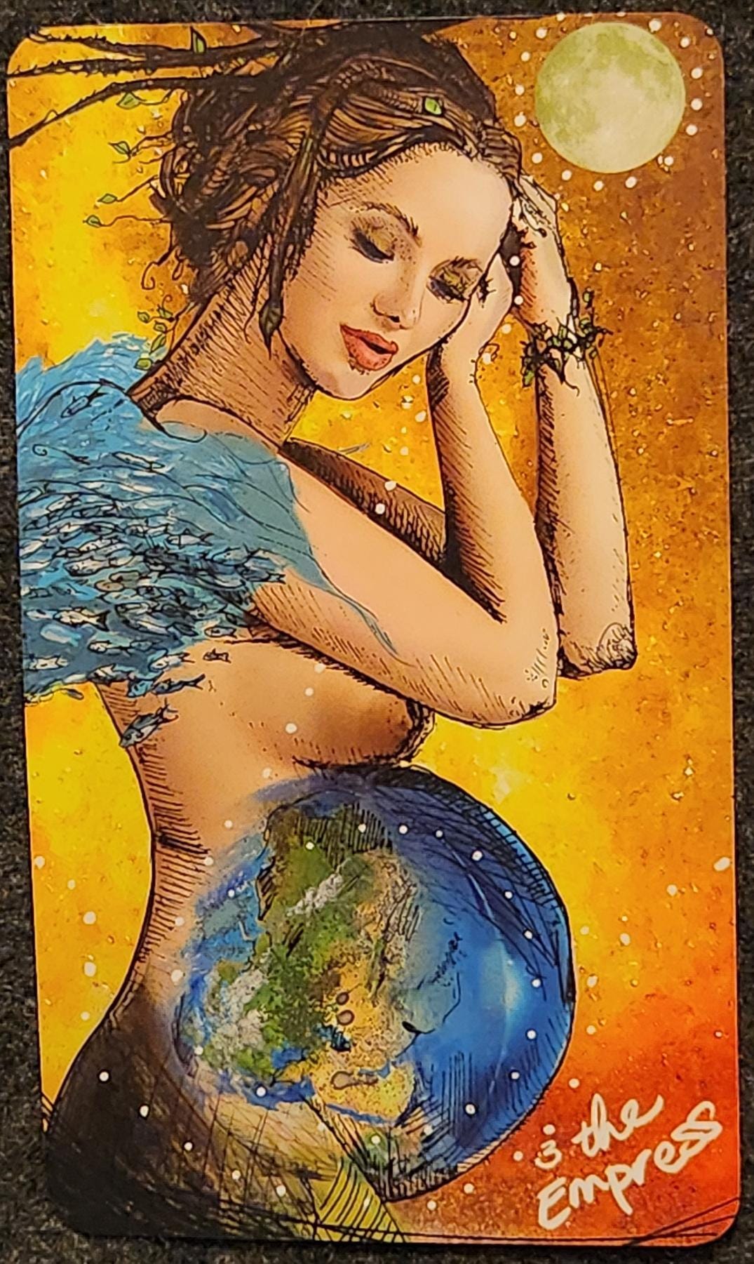 A tarot card labelled 3. The Empress. A white woman who appears pregnant is resting her head on her own hands, her eyes closed. Her pregnant belly resembles Earth, with land masses and oceans. She is topless. Her shoulders are encompassed with what appears to be a stream or river of water, flowing off her back. There is a full moon in the sky above her head. Her hair is in dreadlocks, with vines and leaves woven throughout. 