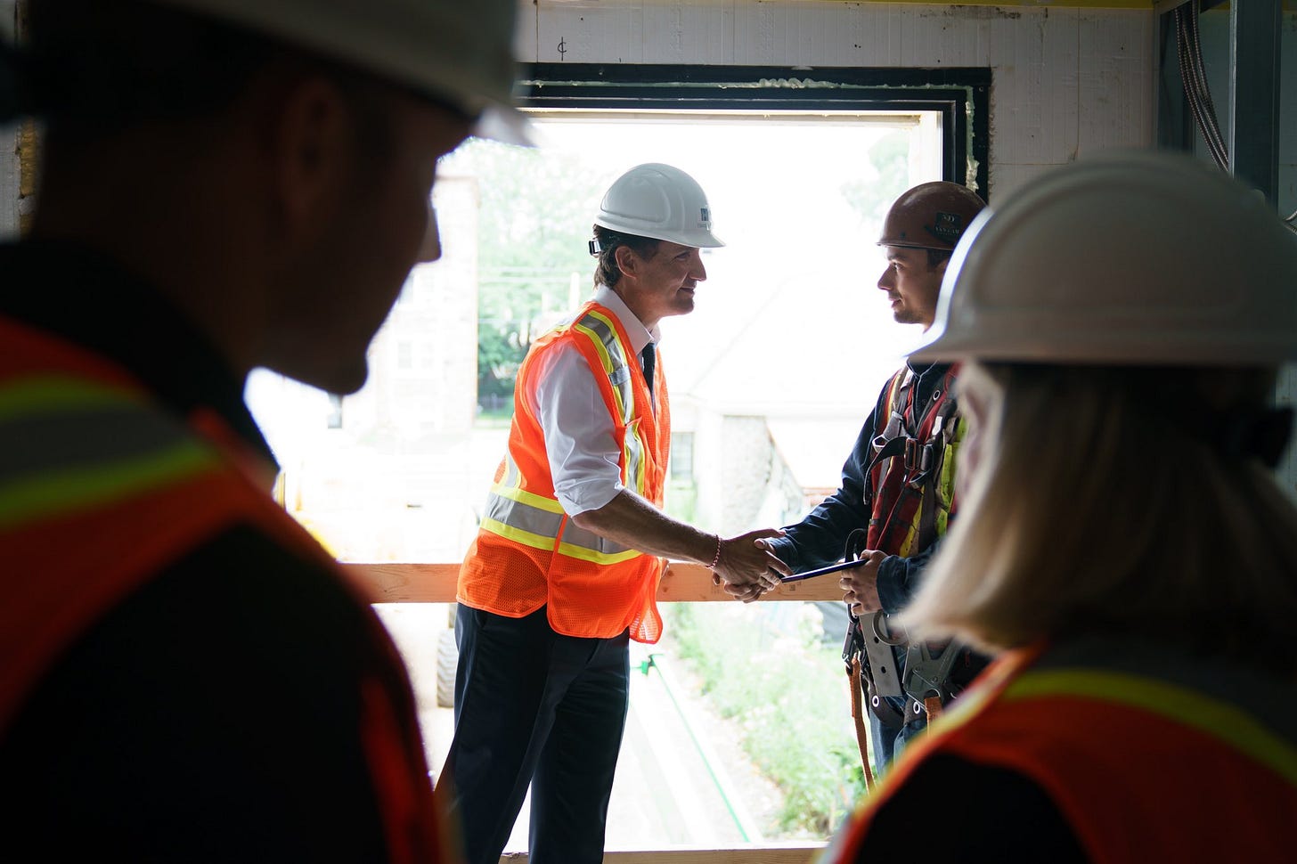 Le premier ministre Justin Trudeau est debout près du cadre d'une fenêtre à l'intérieur d'un chantier de construction. Il serre la main d'un ouvrier. Deux autres personnes sont debout à l'avant-plan. Elles portent des gilets réfléchissants et des casques de protection.