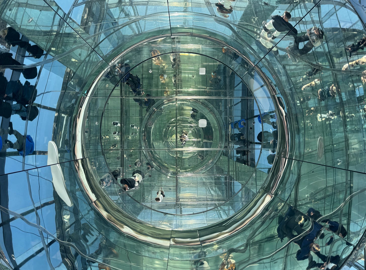 The author’s view looking up through a circular glass opening in a mirrored art installation, with visitors reflected and distorted into infinite recursive forms The author’s view looking up through a circular glass opening in a mirrored art installation, with visitors reflected and distorted into infinite recursive forms