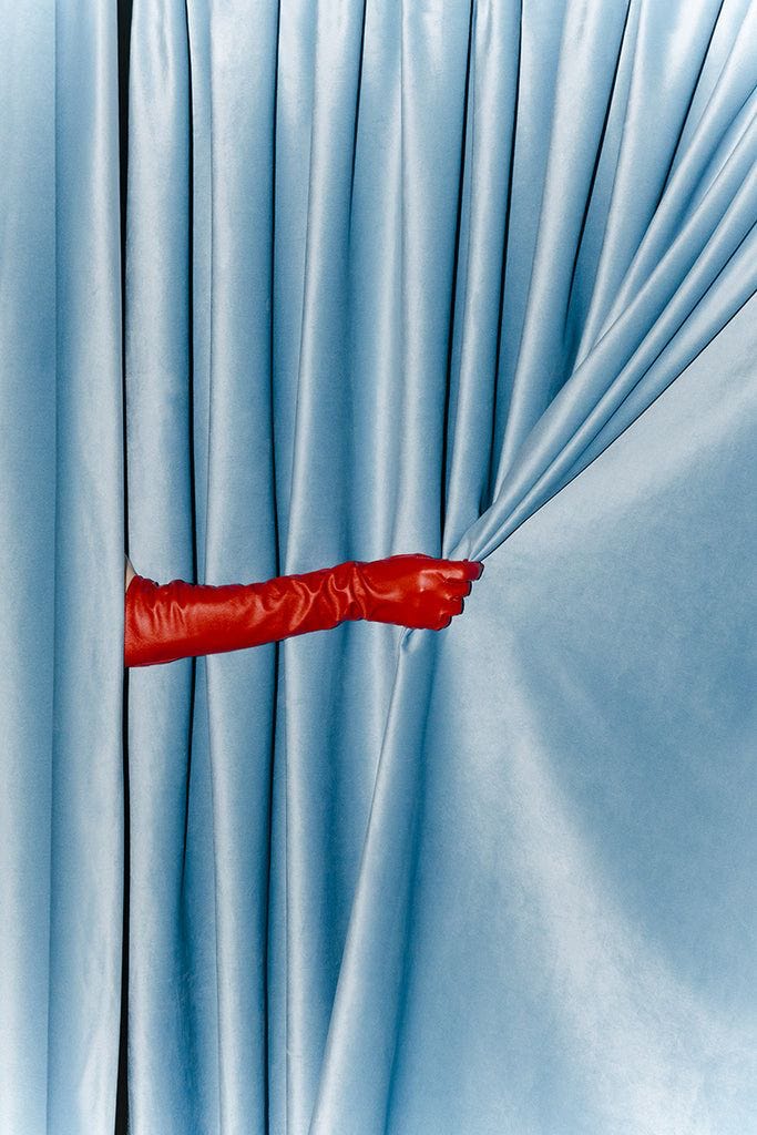 Minimalist fashion photo of a red leather gloved hand pulling back soft blue curtains, bold color contrast, contemporary editorial style