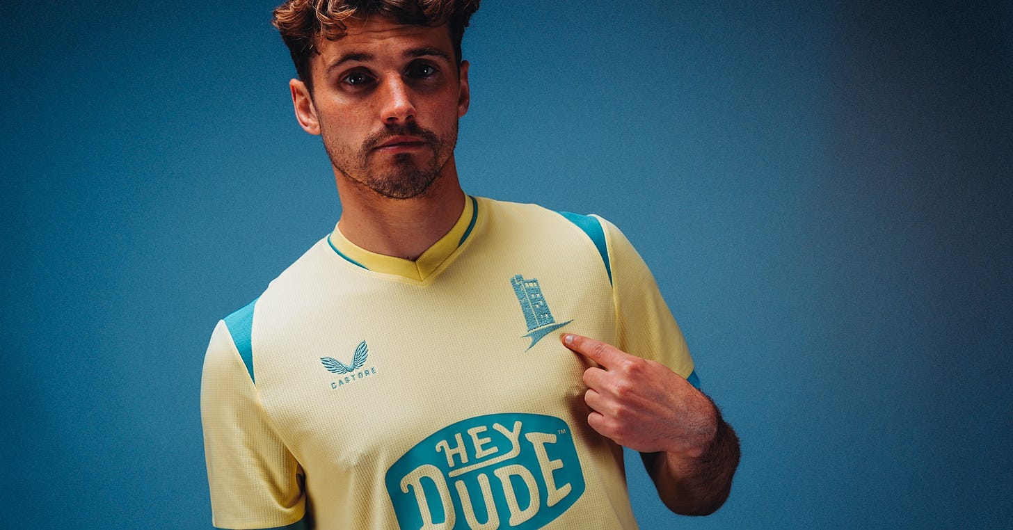 A player models Huddersfield Town’s third kit for the 2026/27 season, a pale yellow shirt with turquoise panelling on the shoulders and collar. He points to the turquoise Victoria Tower crest on the chest, which replaces the usual club badge, with the HeyDude sponsor logo visible below.