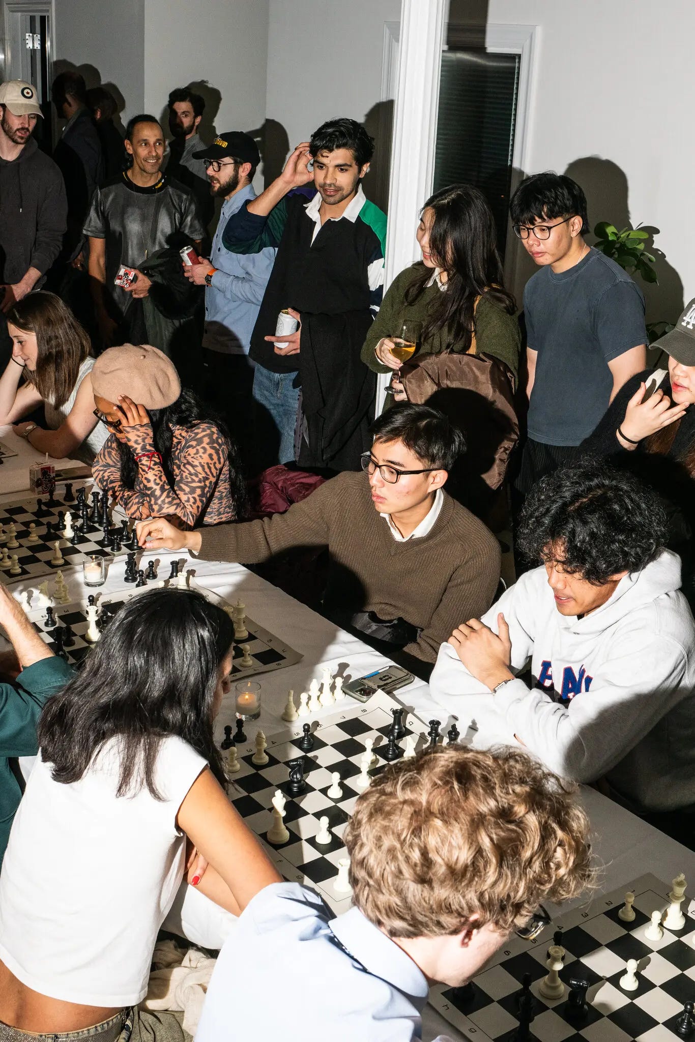 People who appear to be in their 20s and 30s mill about in a white-walled apartment, some holding glasses of wine or cans of beer. others sit at a long table, staring down chessboards, in the middle of games.