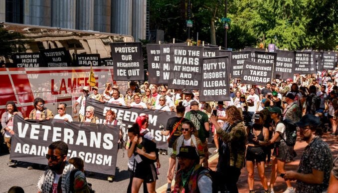 Los manifestantes llevan numerosas pancartas. En la parte superior izquierda se lee «PROTECT OUR NEIGHBORS - ICE OUT» (Protejamos a nuestros vecinos, ¡Fuera el ICE!), y «MILITARY ICE OUT» (¡Fuera el ICE militar!). El texto central dice «WARS AND DISOBEY CONSCIENCE TAKES COURAGE» (Las guerras y desobedecer: la conciencia requiere valentía" Los manifestantes llevan numerosas pancartas. En la parte superior izquierda se lee «PROTECT OUR NEIGHBORS - ICE OUT» (Protejamos a nuestros vecinos, ¡Fuera el ICE!), y «MILITARY ICE OUT» (¡Fuera el ICE militar!). El texto central dice «WARS AND DISOBEY CONSCIENCE TAKES COURAGE» (Las guerras y desobedecer: la conciencia requiere valentía"