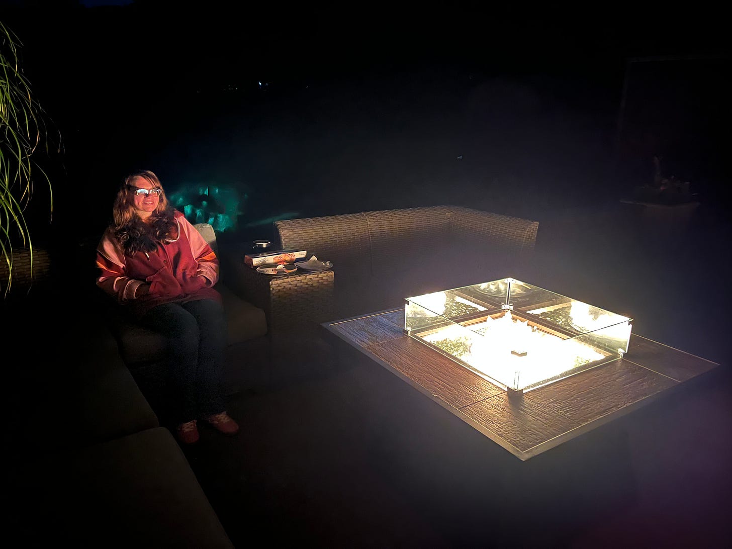 Woman sitting in the dark in front of a gas firepit with a green glowing thing just over her left shoulder.