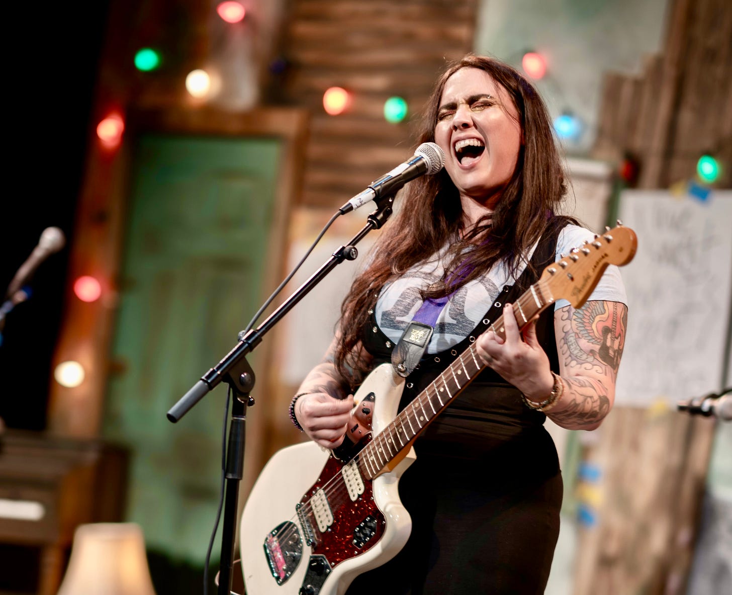 A heavily tattooed young wman rocks out on guitar while belting into a microphone