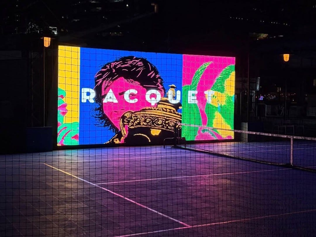 A tennis court at night at Racquet Club at the Seaport, with an illuminated RACQUET screen and the New York skyline beyond. A tennis court at night at Racquet Club at the Seaport, with an illuminated RACQUET screen and the New York skyline beyond.