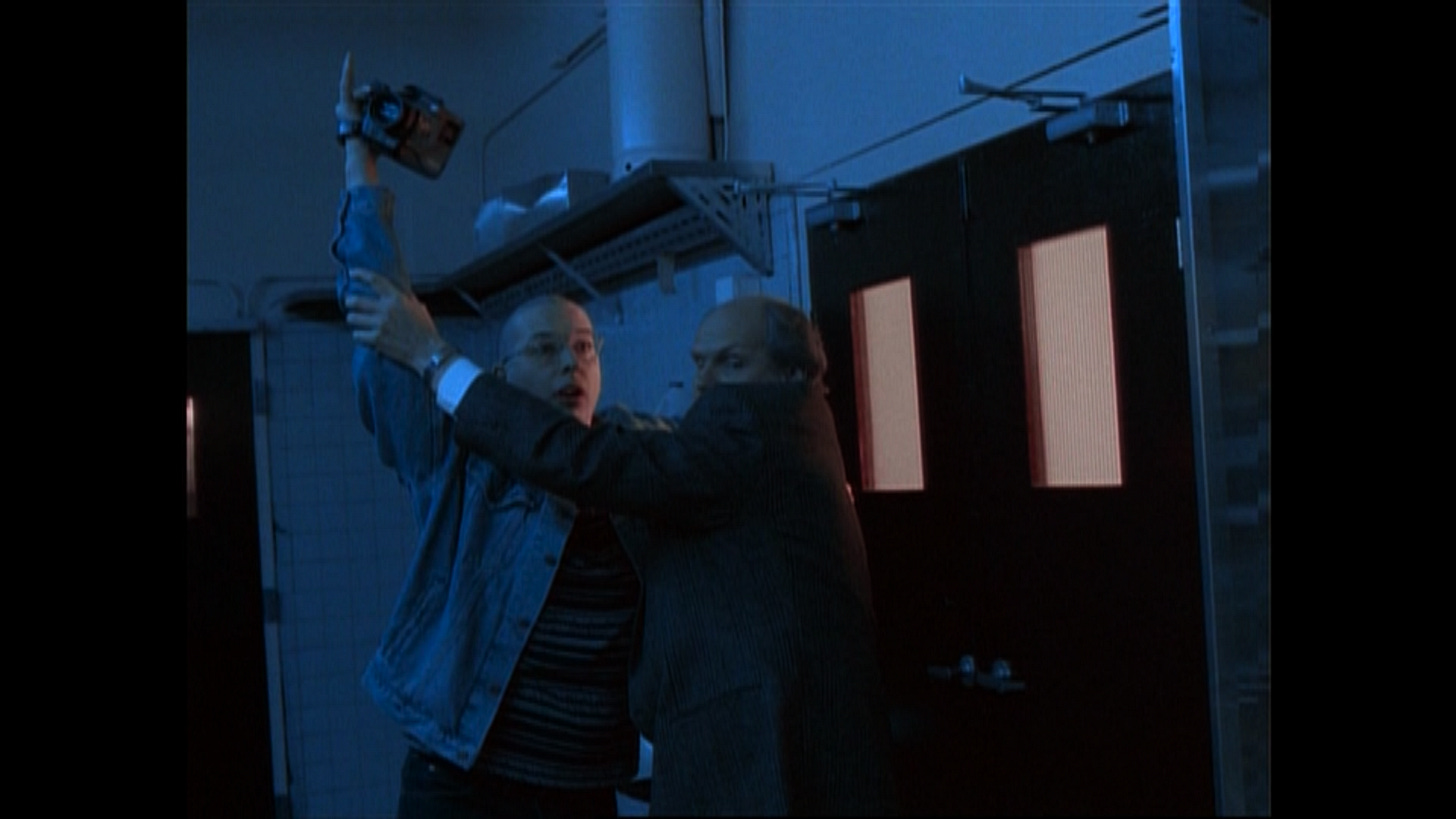 Detective Manners fights to prevent Blaine from filming the alien autopsy. Blaine is holding a very large, 90s style camcorder. Detective Manners fights to prevent Blaine from filming the alien autopsy. Blaine is holding a very large, 90s style camcorder.