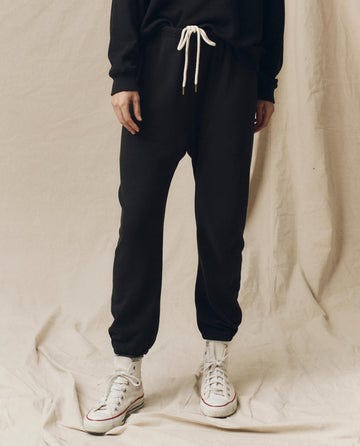 A person in The Stadium Sweatpant in ALMOST BLACK by THE GREAT. CORE KNITS, featuring a white drawstring, and white high-top sneakers stands on a beige background.