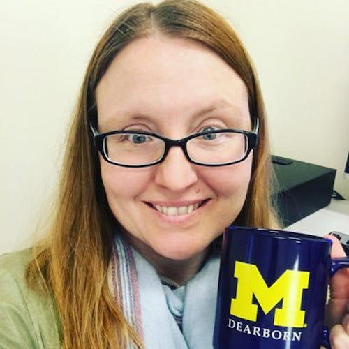 A head-and-shoulders portrait shows a person with long light-brown hair wearing glasses and smiling at the camera while holding a dark blue mug with a large yellow “M” and the word “Dearborn.” The person wears a light scarf and appears indoors in an office-like environment with a neutral background and desk items partially visible.