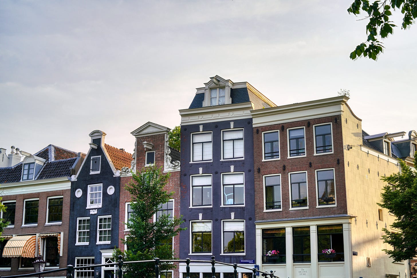 Row of historic narrow Amsterdam canal houses with varied brick and painted facades, gabled roofs, and tall windows beneath a pale sky. Row of historic narrow Amsterdam canal houses with varied brick and painted facades, gabled roofs, and tall windows beneath a pale sky.