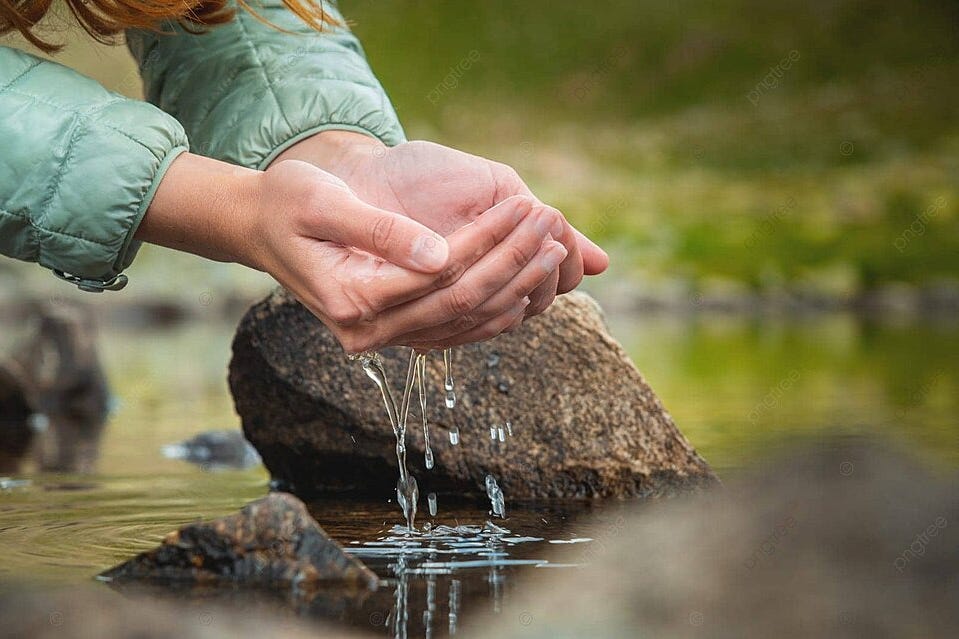 Hands cupped, collecting water from a stream. Person wearing a light green jacket. Rocky and grassy background, serene mood.