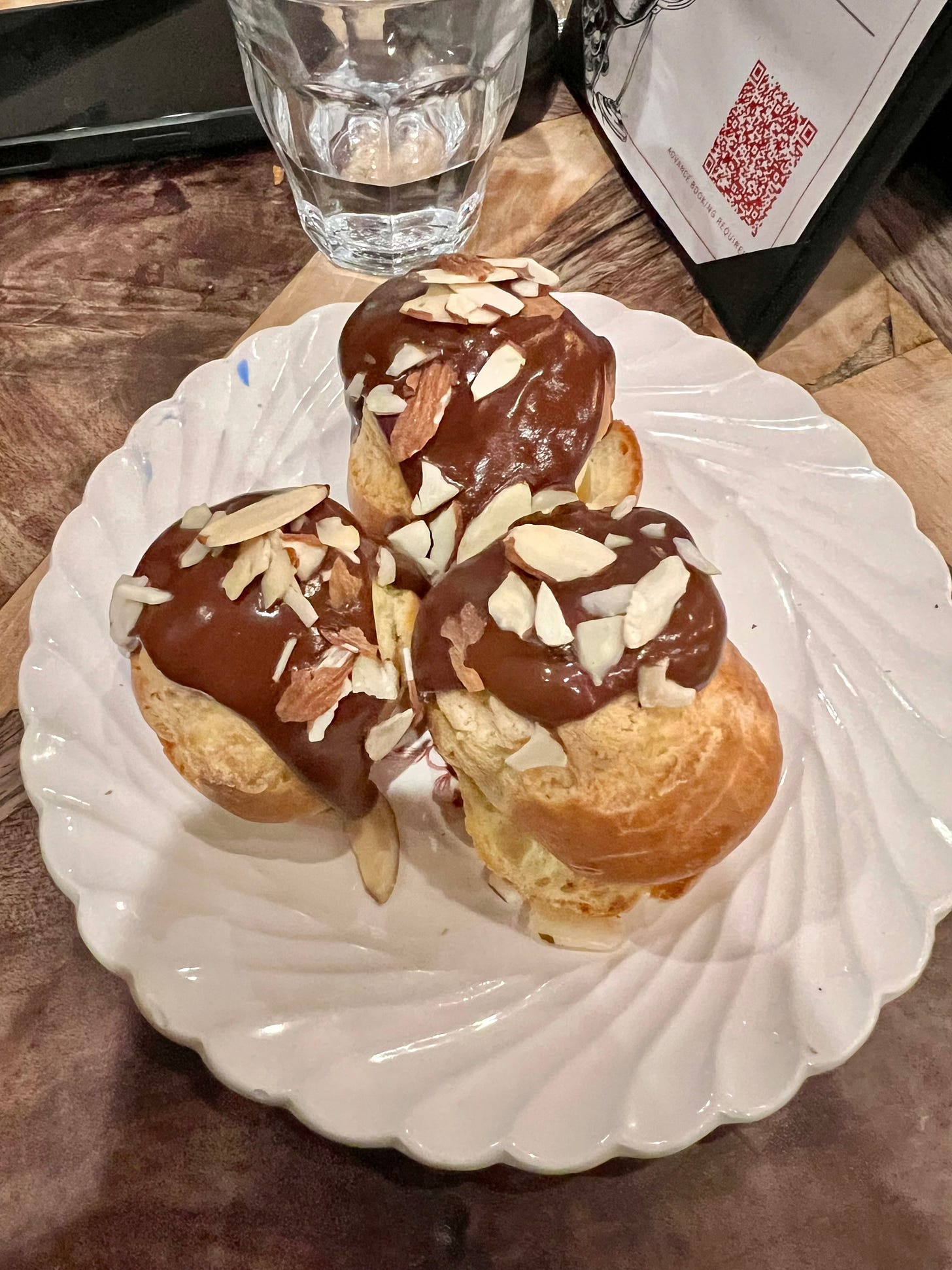 Three profiteroles topped with dark chocolate ganache and sliced toasted almonds, served on a scalloped white plate at Bouillon by Moulin.