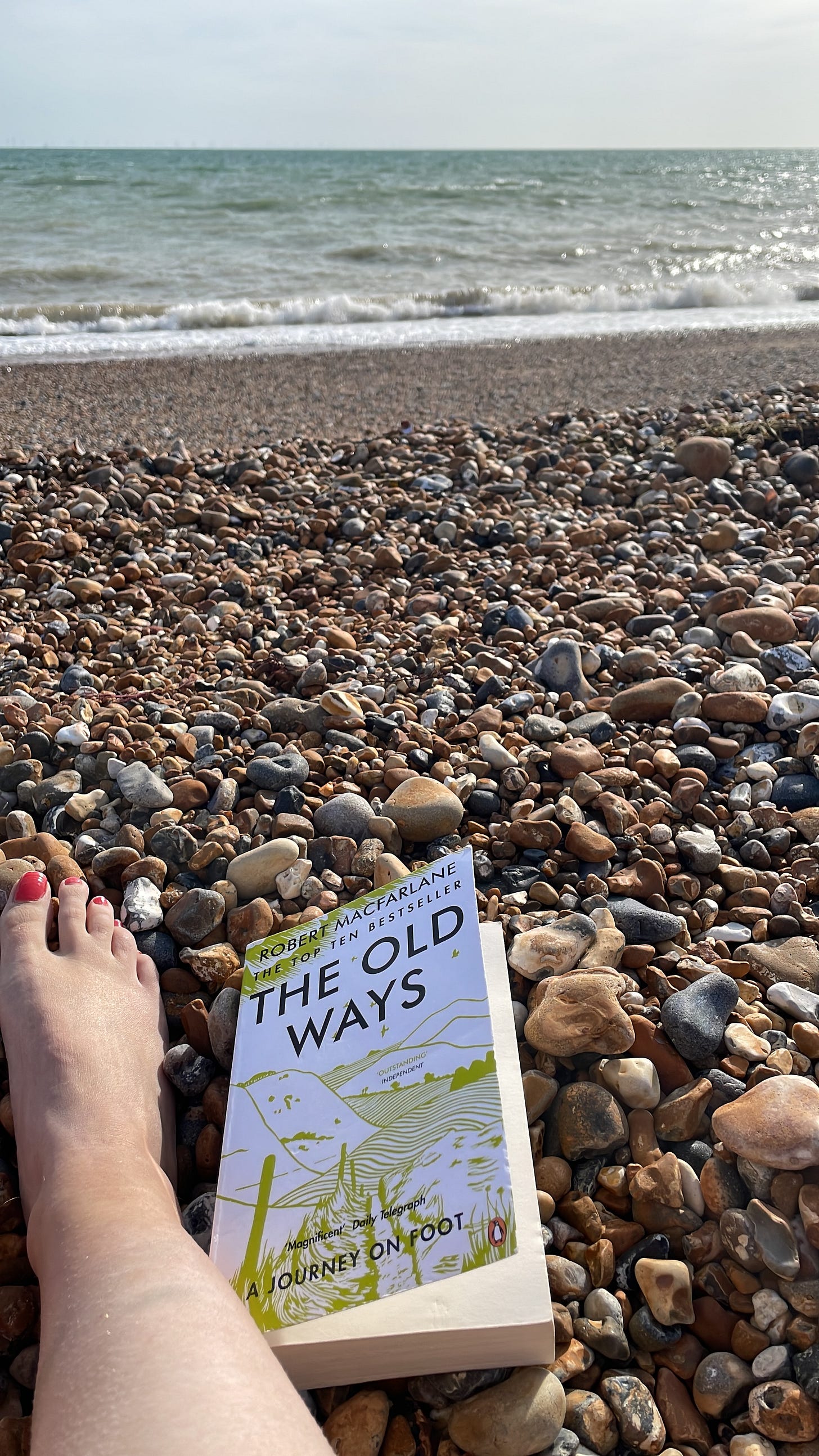Image of a foot next to Robert Macfarlane's 'The Old Ways' on a pebble beach