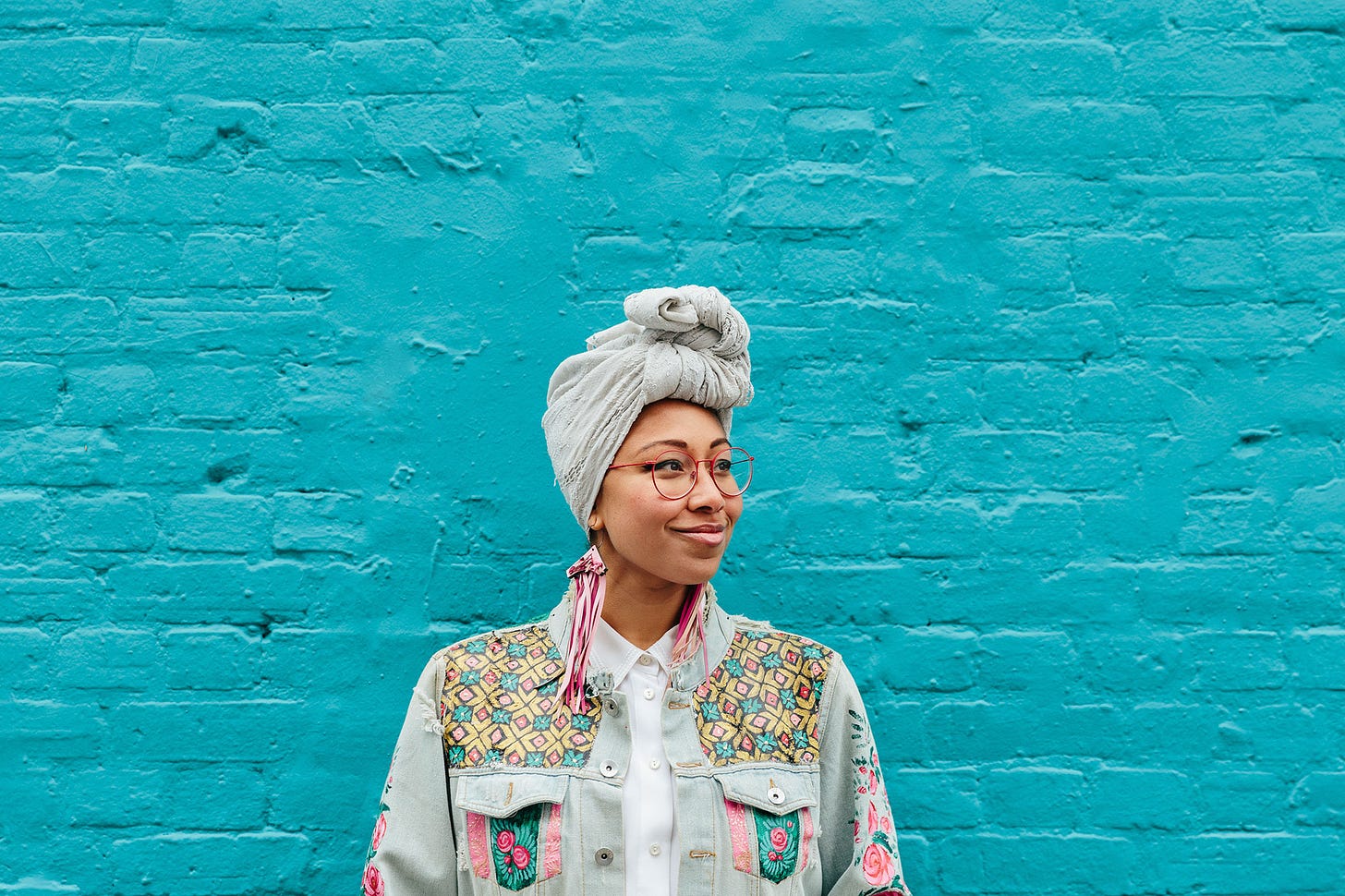 Yassmin Abdel-Magied against a teal brick wall, smiling and looking out to her left. She is in a grey turban, pink long earrings, and a painted jean jacket. The photo is cropped at her torso.