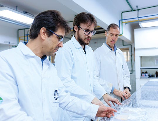 A imagem mostra três homens em um laboratório, todos usando jalecos brancos. Eles estão lado a lado, inclinados sobre uma bancada, observando e manuseando materiais organizados em recipientes ou placas transparentes. Dois deles usam óculos, e todos parecem concentrados na atividade, enquanto o ambiente ao redor apresenta equipamentos e tubulações típicos de laboratório.