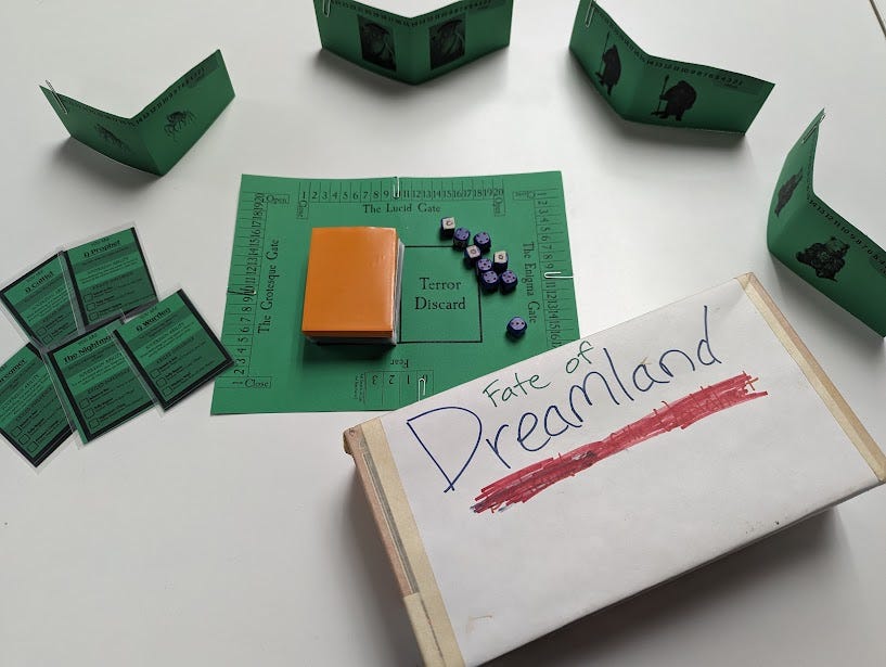 A plain white box with marker "Fate of Dreamland" sharpied on the front. Green paper pieces with paper clip markers are assembled on the table to show how the game would be laid out to play, but this is clearly a prototype.