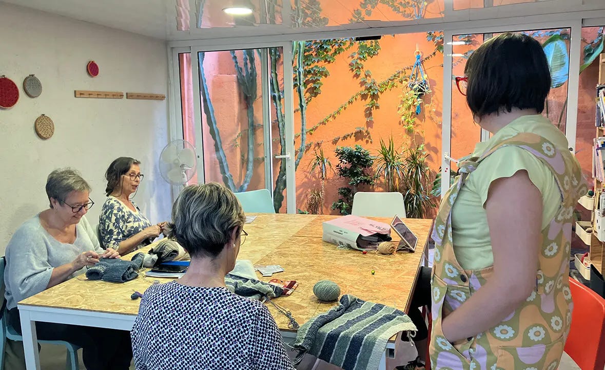 a woman standing and three women sitting by a table knitting