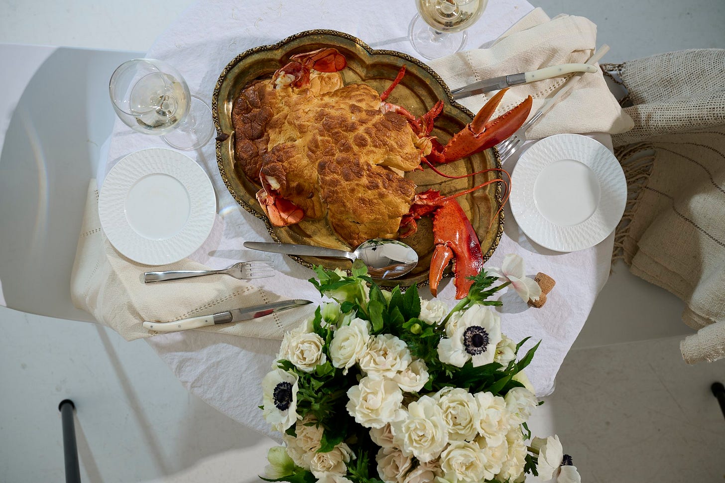 Whole lobster soufflé presented on a silver tray with lobster tails and claws, set on an elegant white table with champagne, white plates, and a bouquet of ivory and anemone flowers.