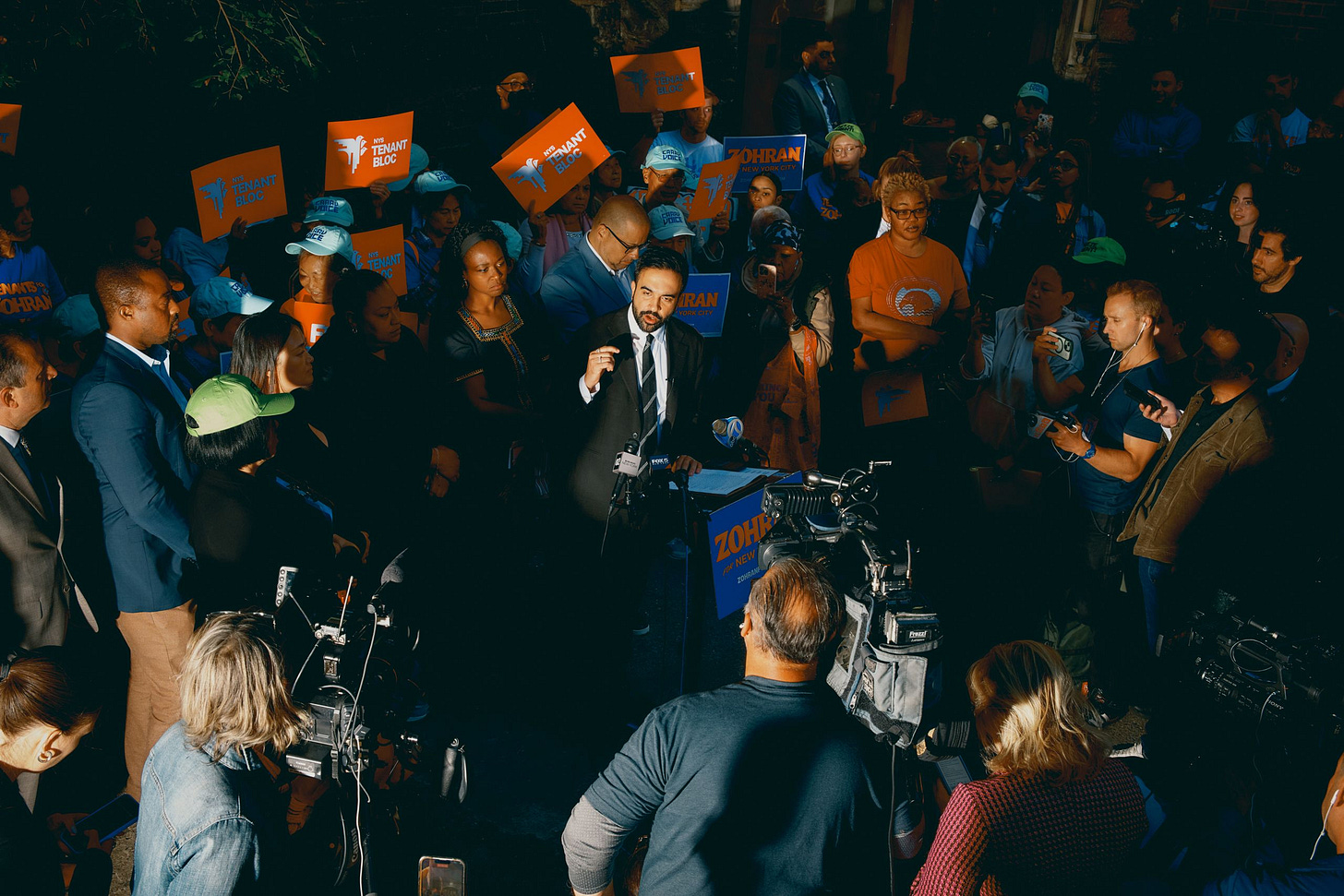 A man in a suit speaks to a crowd of supporters and media.