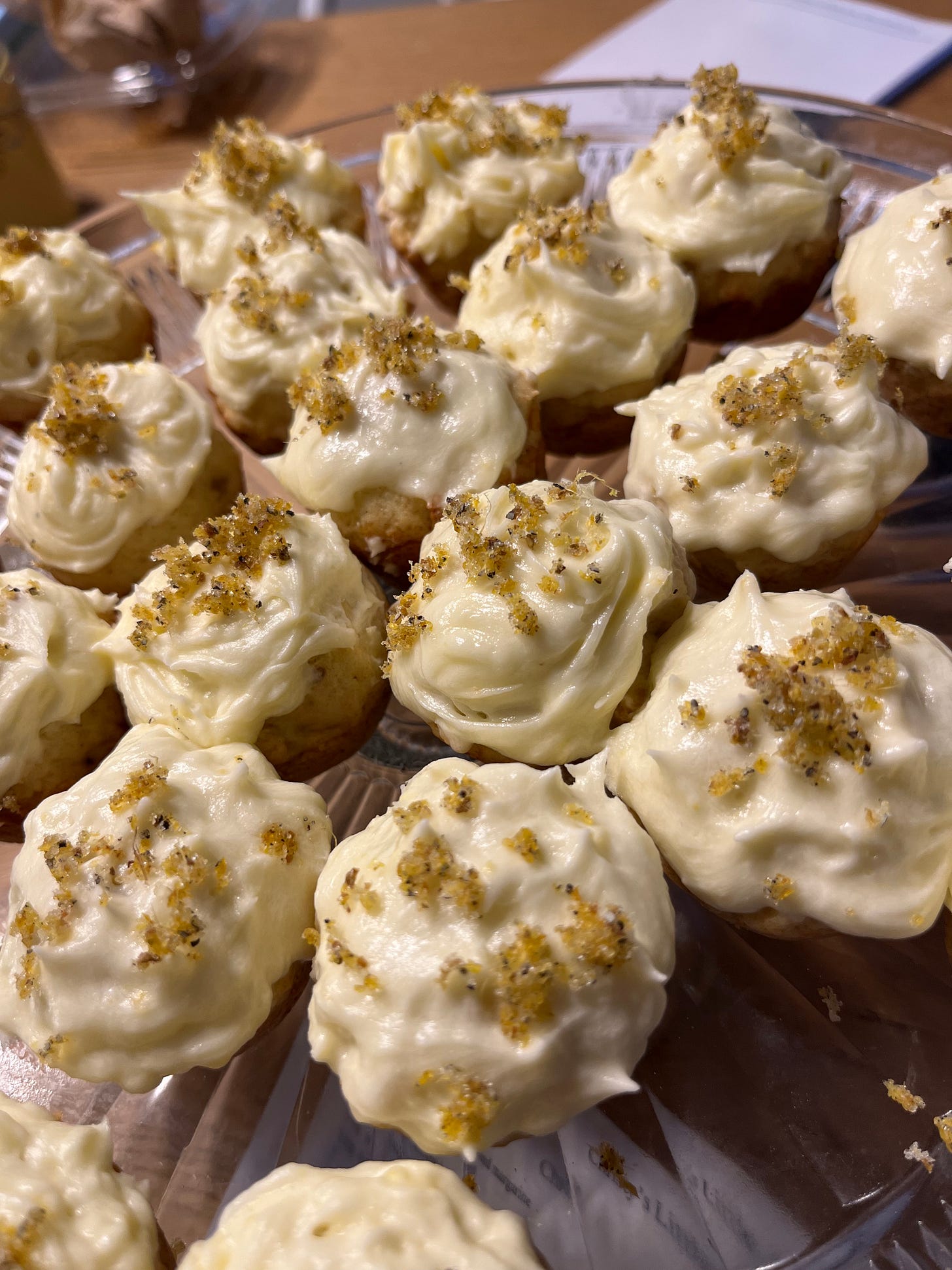 A close-up photograph of mini muffins with pale yellow frosting and sparkly gold and black sprinkled toppings. A close-up photograph of mini muffins with pale yellow frosting and sparkly gold and black sprinkled toppings.