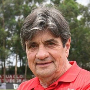 Homem de cabelos grisalhos, olha para a câmera com leve sorriso em ambiente externo. Ele veste camisa polo vermelha, com árvores desfocadas ao fundo. Homem de cabelos grisalhos, olha para a câmera com leve sorriso em ambiente externo. Ele veste camisa polo vermelha, com árvores desfocadas ao fundo.