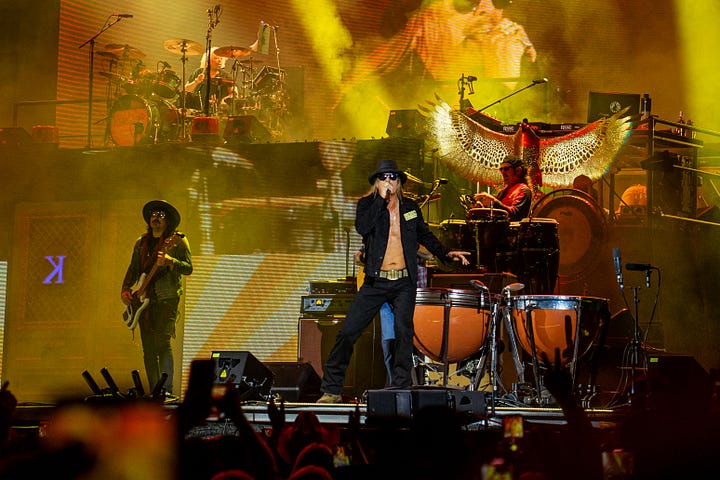 Kid rock is wearing a black open face button down, black hat and black sun glasses singing on stage with his band while the crowd dances to his music