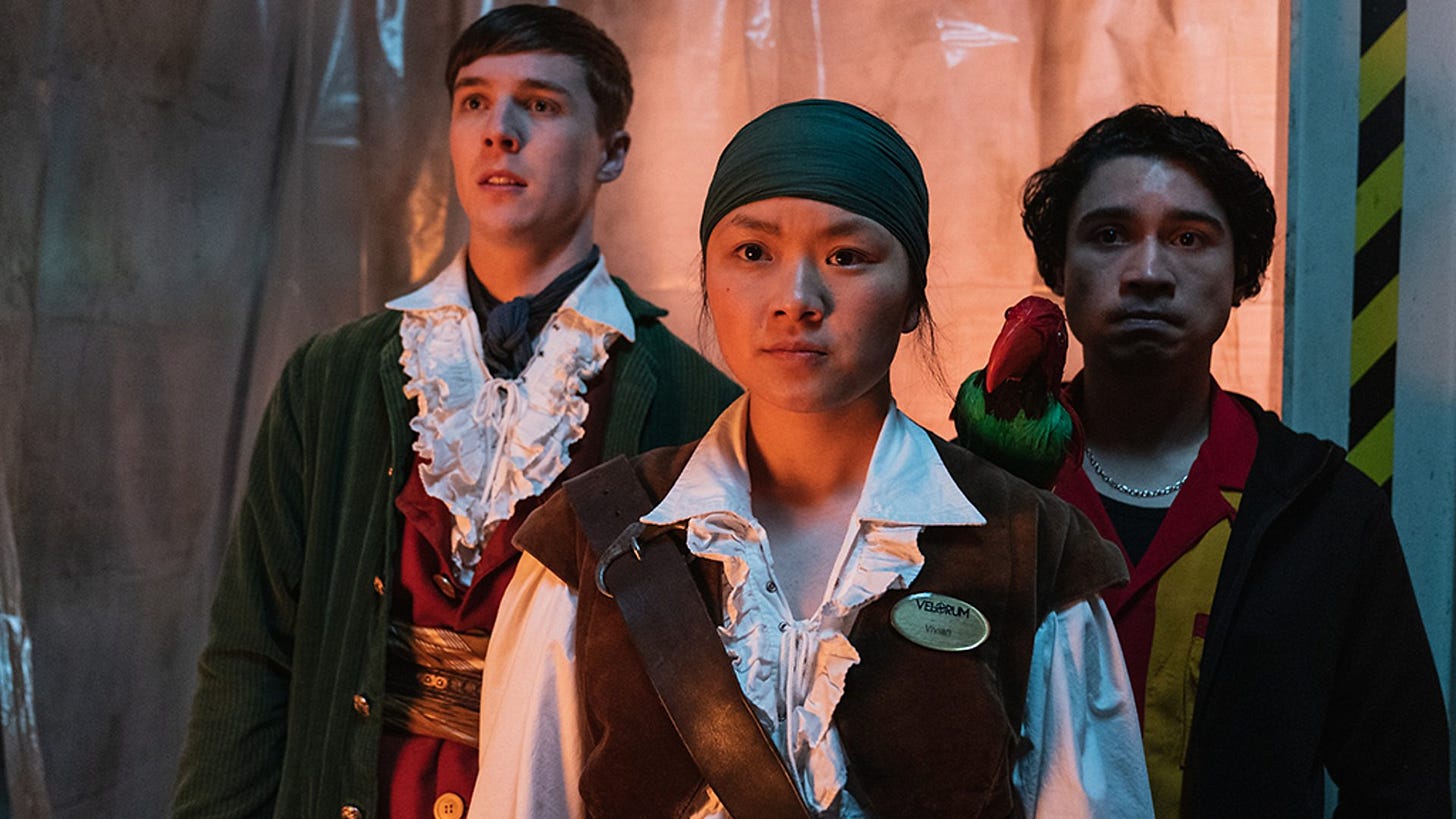 Three people are standing looking slightly off camera, all wearing costumey pirate garb. They are in an industrial setting, with clear plastic curtain and yellow-and-black safety tape on a white wall.