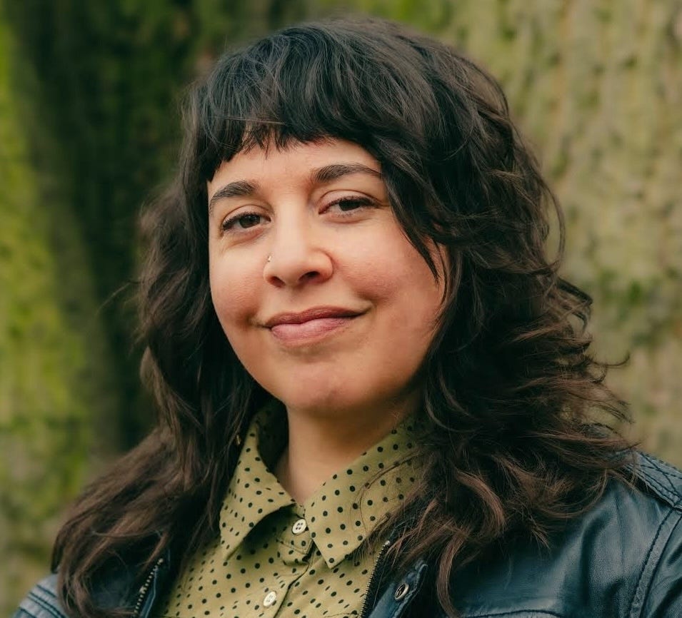 headshot photo of Iris. She is looking directly at the camera with a half smile, long dark hair in waves, a jean jacket and collared shirt. Light skin. With a forest background.