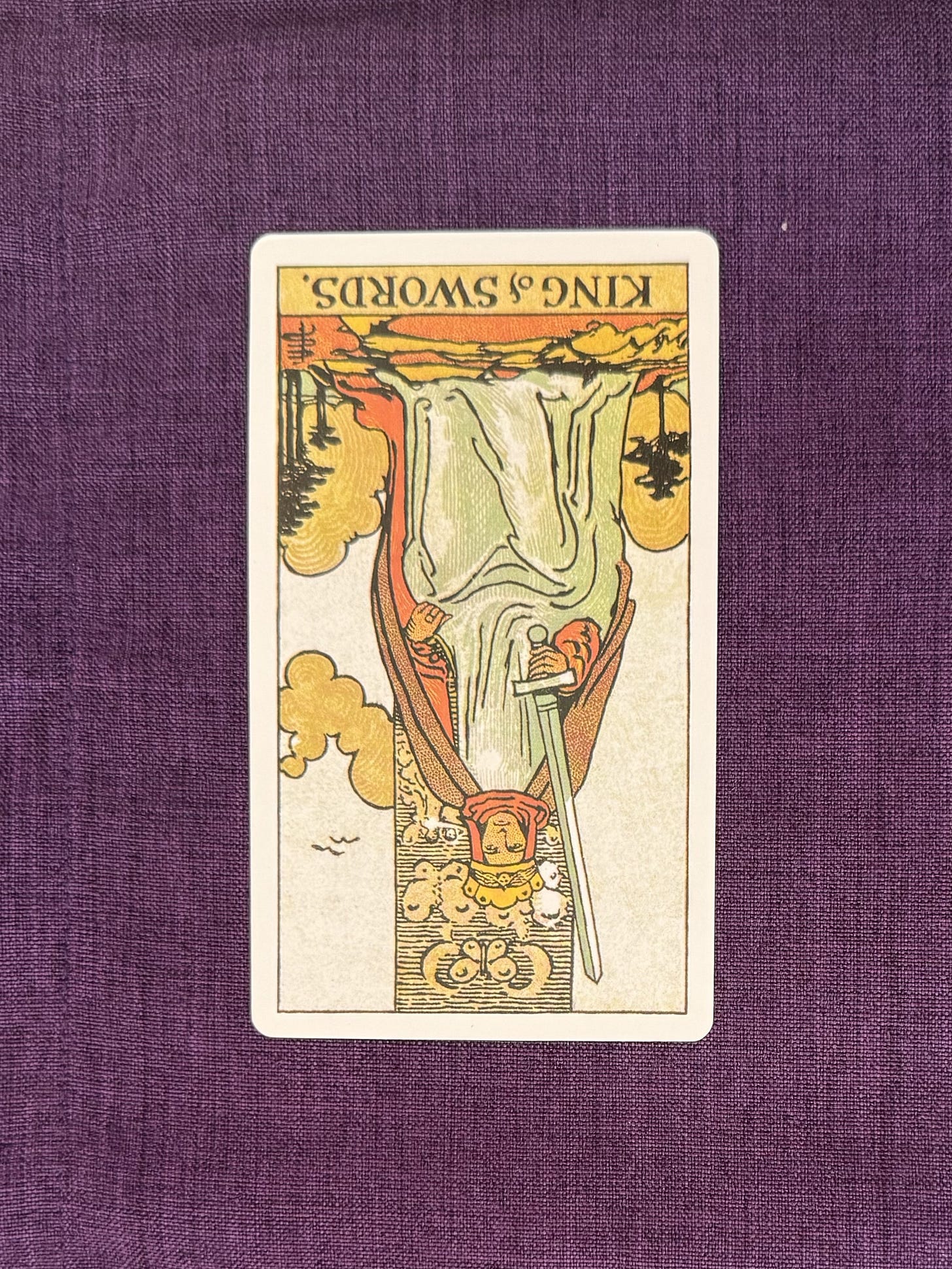 A seated figure sits on a stone throne set against a clear sky with light clouds. The figure wears a crown and a long robe, and holds an upright sword in one hand, positioned vertically. The other hand rests on the arm of the throne. The throne is decorated with carved motifs and has a high back. The figure faces forward, with wings and small carved figures visible on the throne. The ground beneath is flat and bare.