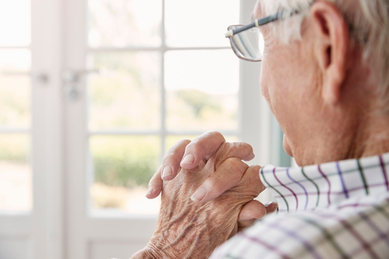 Tight side view of older white man with glasses and white hair. He's home, looking out the window, with hands folded against his shoulder.
