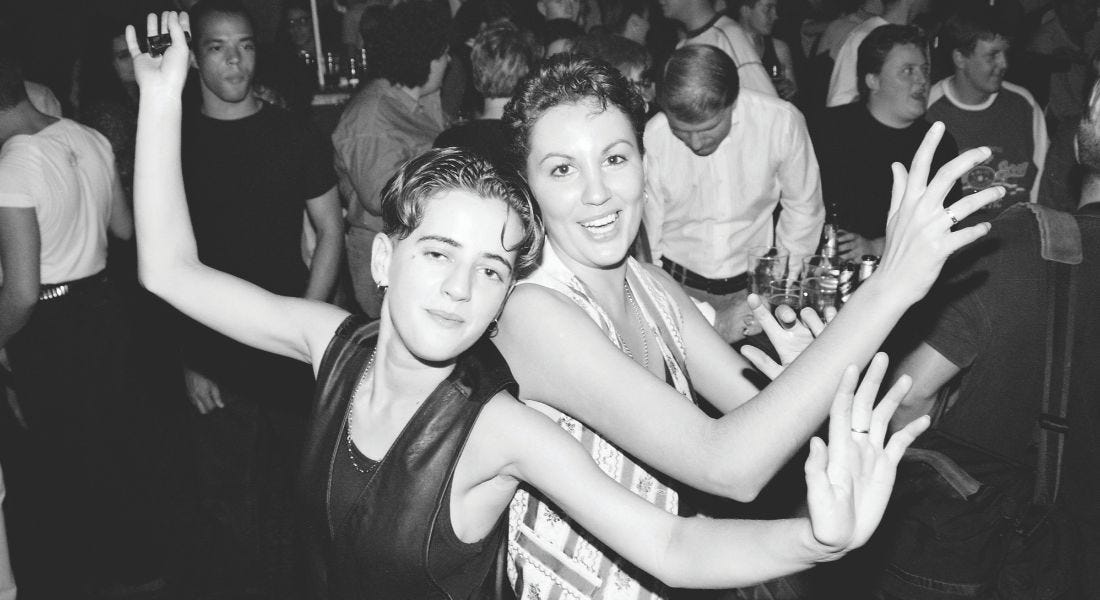 A black and white picture of a couple smiling into the camera dancing together, with a crowd of people behind them A black and white picture of a couple smiling into the camera dancing together, with a crowd of people behind them