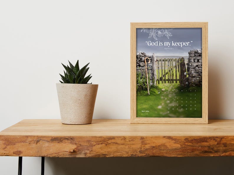 May include: A framed print featuring a pastoral scene with a stone gate and a path leading to it, with the quote 'God is my keeper.' The print is displayed on a wooden surface next to a potted succulent plant. The frame is light-colored, and the print has a calendar for May 2024.