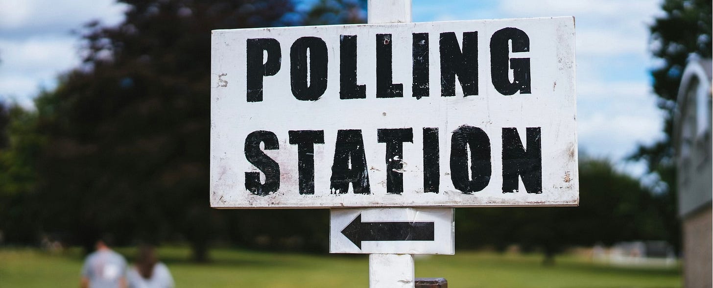 large white sign with black arrow and words Poling Station. Sign is against background of a field and 2 people walking away.