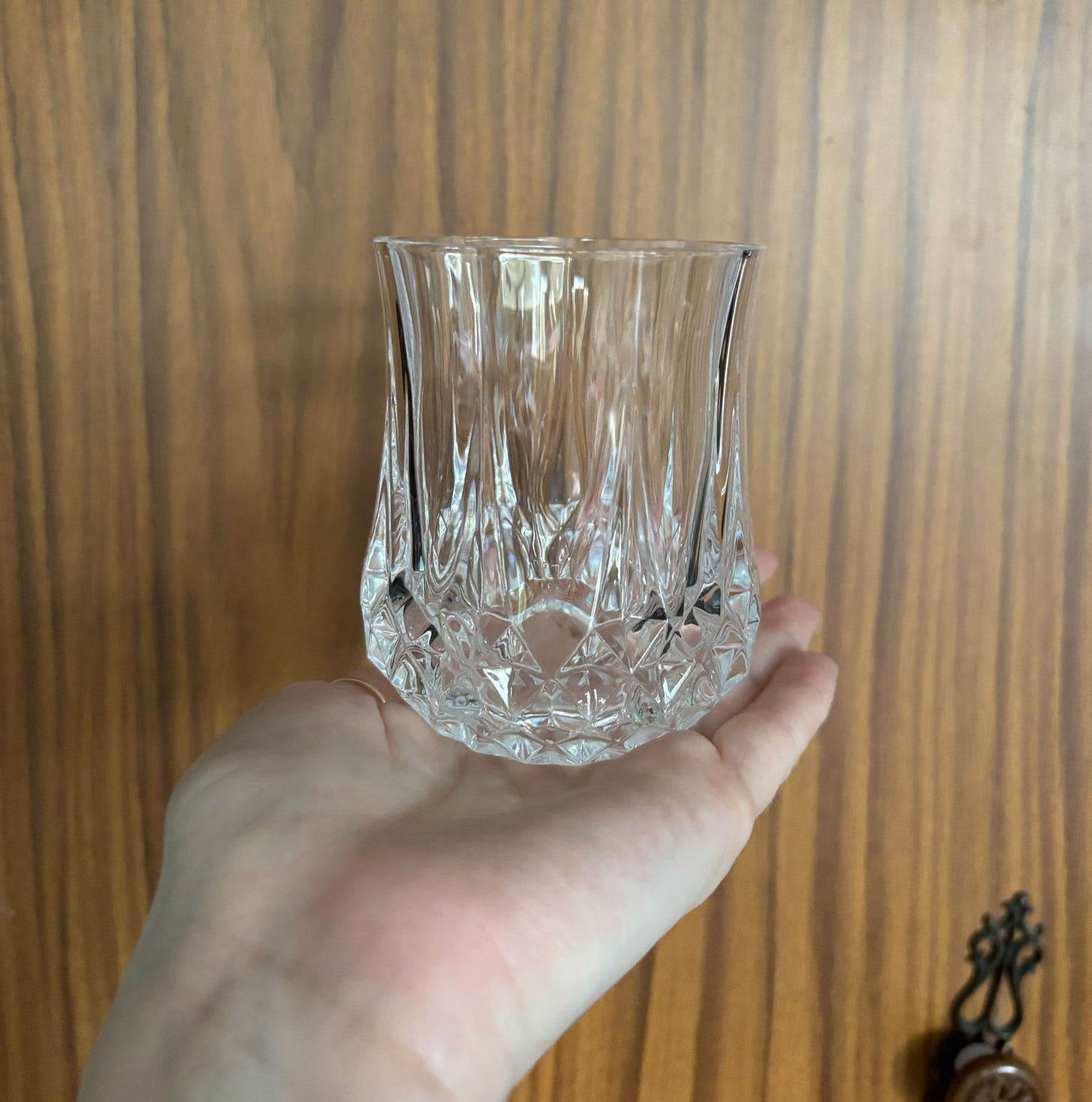An extended hand holds a small cut glass tumbler infant of an old woodgrain wardrobe