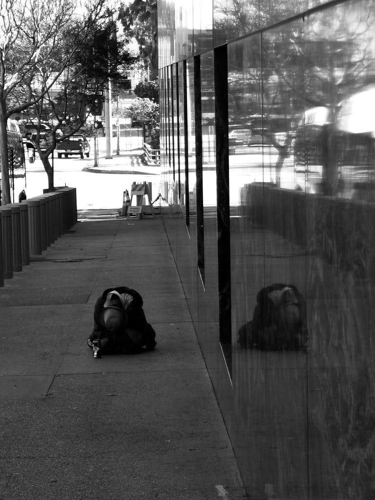 Monochrome photo of a homeless man seated, slumped over, on a sidewalk in downtown Los Angeles. The sidewalk has concrete bollards on the street side and a poliched concrete wall on the other side, in which the man is reflected. A sunlit street is shown in the background.