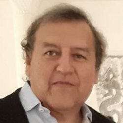 Um homem de meia-idade com pele clara, cabelos curtos e ondulados, parcialmente grisalhos, e está com expressão neutra. Veste uma camisa azul-clara e um blazer preto