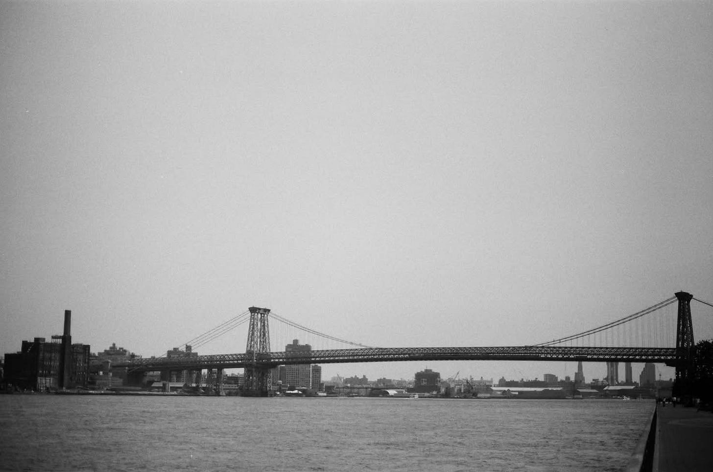 A grainy, black-and-white film photograph of the Williamsburg Bridge stretching across the East River in New York City. The massive steel suspension bridge spans the horizon, connecting industrial waterfront buildings and a distant city skyline under a vast, overcast gray sky. A grainy, black-and-white film photograph of the Williamsburg Bridge stretching across the East River in New York City. The massive steel suspension bridge spans the horizon, connecting industrial waterfront buildings and a distant city skyline under a vast, overcast gray sky.