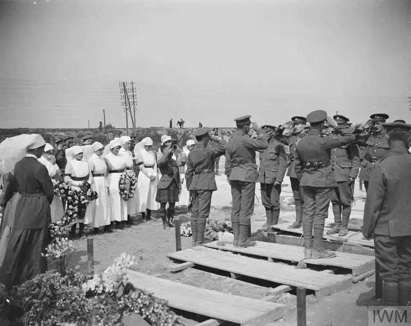 The image depicts a solemn scene of military personnel saluting at a gravesite, with women in white dresses and hats holding wreaths standing nearby. The soldiers are dressed in uniforms and caps, standing on wooden planks. In the foreground, flowers and plants are visible. A watermark "IWM" is present in the bottom-right corner. The image depicts a solemn scene of military personnel saluting at a gravesite, with women in white dresses and hats holding wreaths standing nearby. The soldiers are dressed in uniforms and caps, standing on wooden planks. In the foreground, flowers and plants are visible. A watermark "IWM" is present in the bottom-right corner.