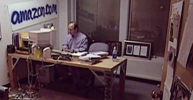 Jeff Bezos in 1999 sitting at a desk in the first Amazon office with a hand-painted "amazon.com" sign on the wall