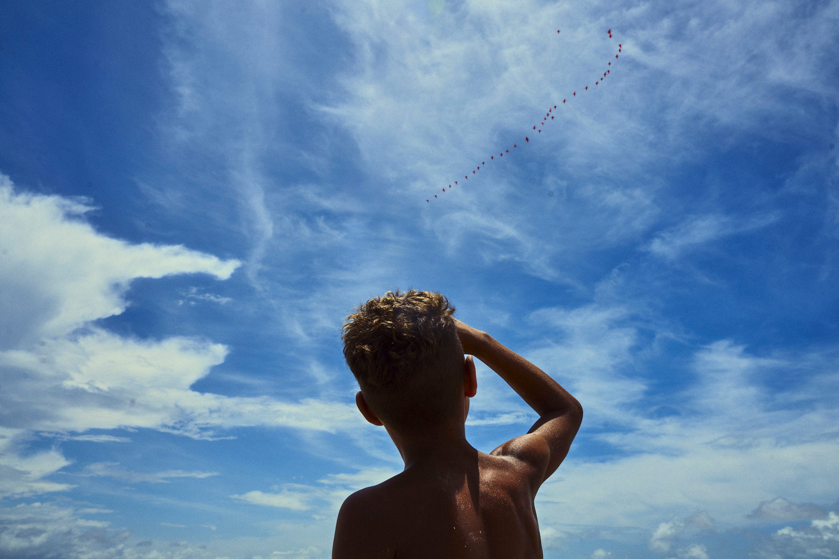 A child with short, curly hair stands shirtless, looking up at a long string of red kites flying high in a partly cloudy blue sky. The child raises a hand to shield their eyes from the sun.