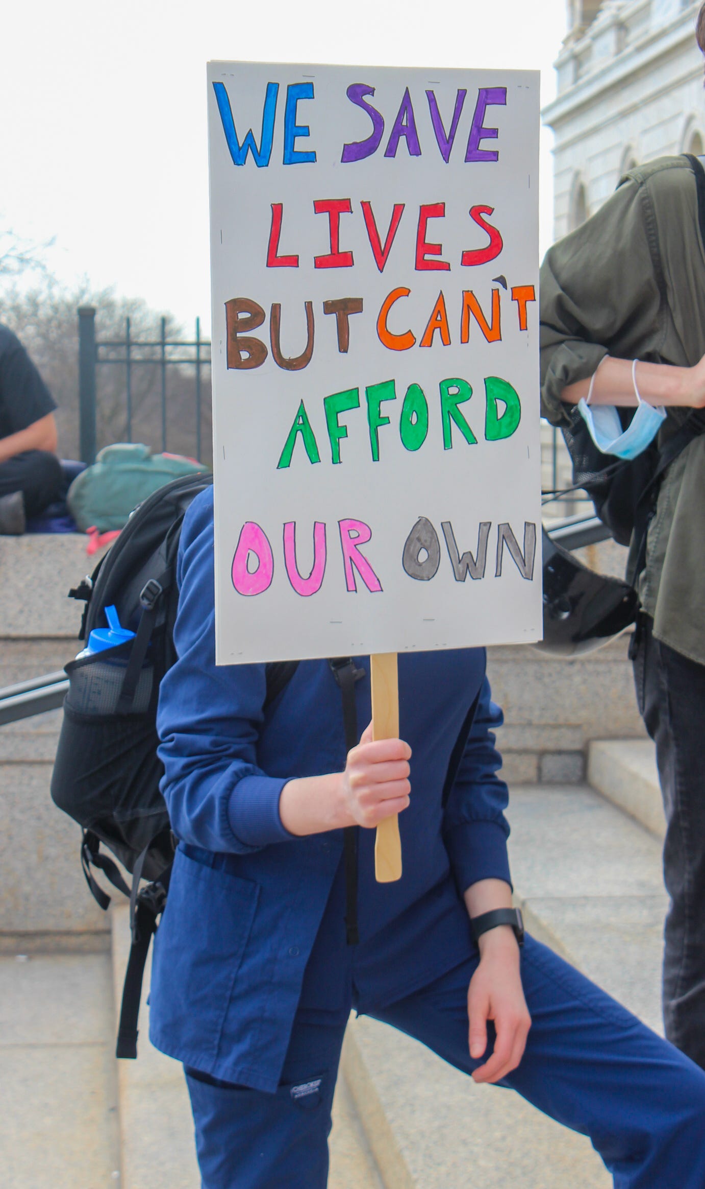 a healthcare worker in blue scrubs holds a sign in rainbow letters reading "we save lives but can't afford our own"