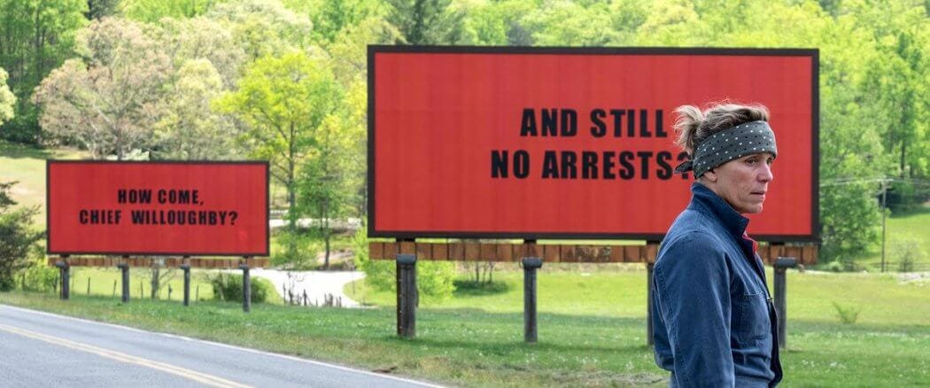 Shot from the movie Three Billboards. One bill board in red behind Frances McDormand reads "And still no arrests." The red billboard further back reads "How come, Chief Willoughby?"
