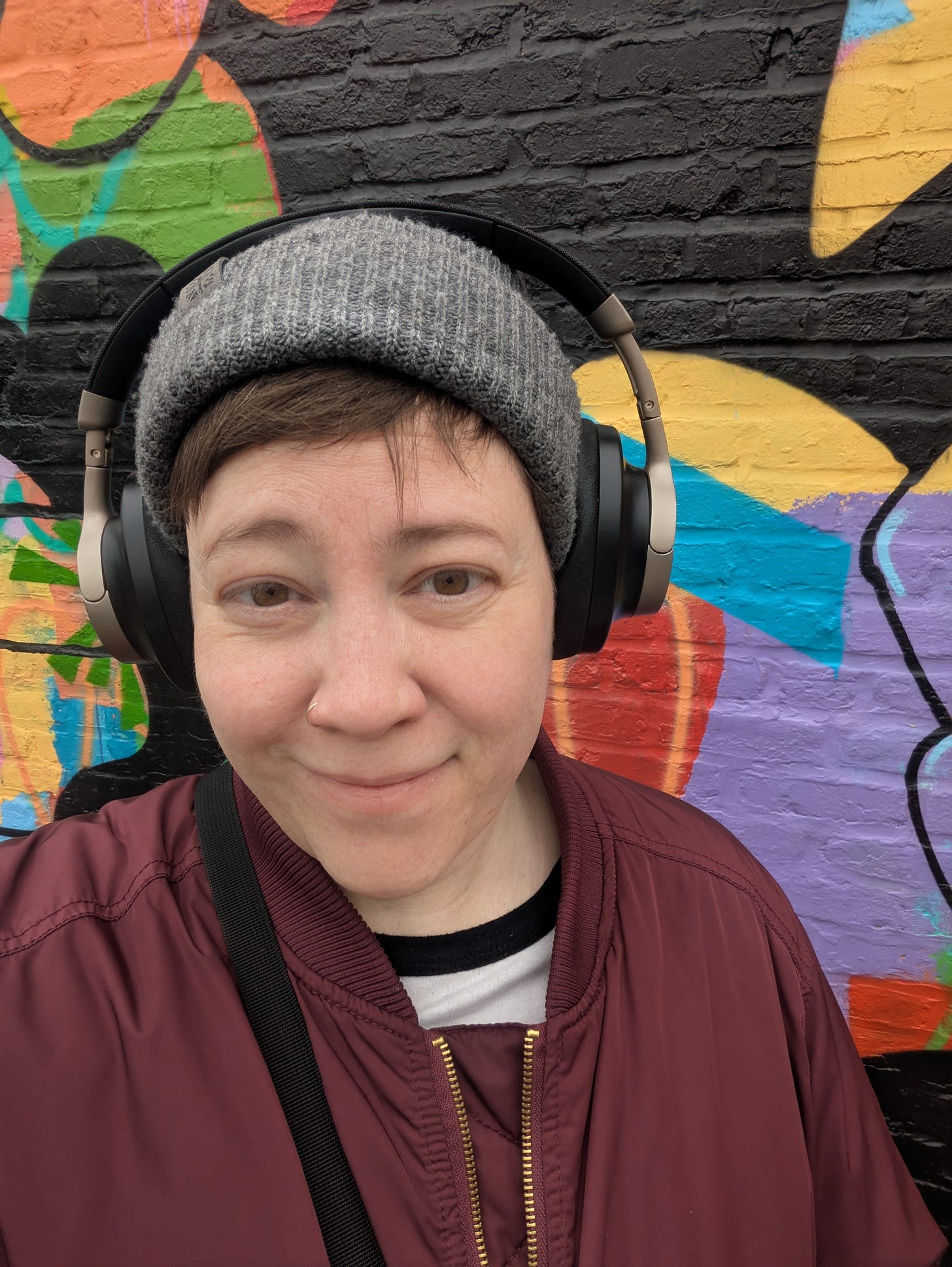 White woman with a slight smile wearing a gray knit cap, maroon jacket, and over the ear wireless headphones standing against a colorful graffitied brick wall. 