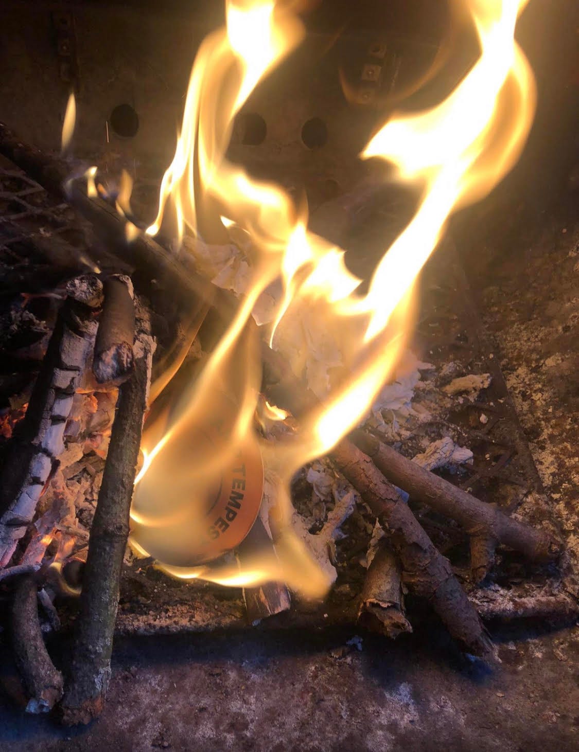 A small fire in a metal container. There is a pile of sticks and flames and a disposable branded coaster in the flames. A small fire in a metal container. There is a pile of sticks and flames and a disposable branded coaster in the flames.