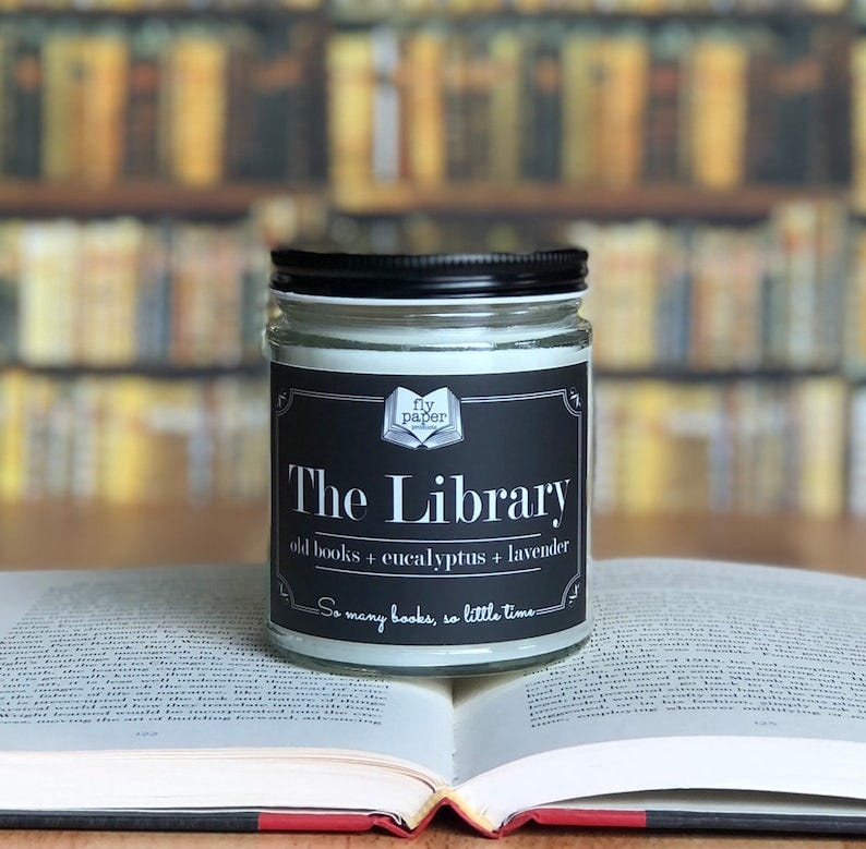 May include: A black candle jar with a white label that reads 'The Library' and 'old books + eucalyptus + lavender' in black text. The label also includes the text 'fly paper' and 'So many books, so little time' in smaller black text. The candle jar is sitting on an open book with a white background.