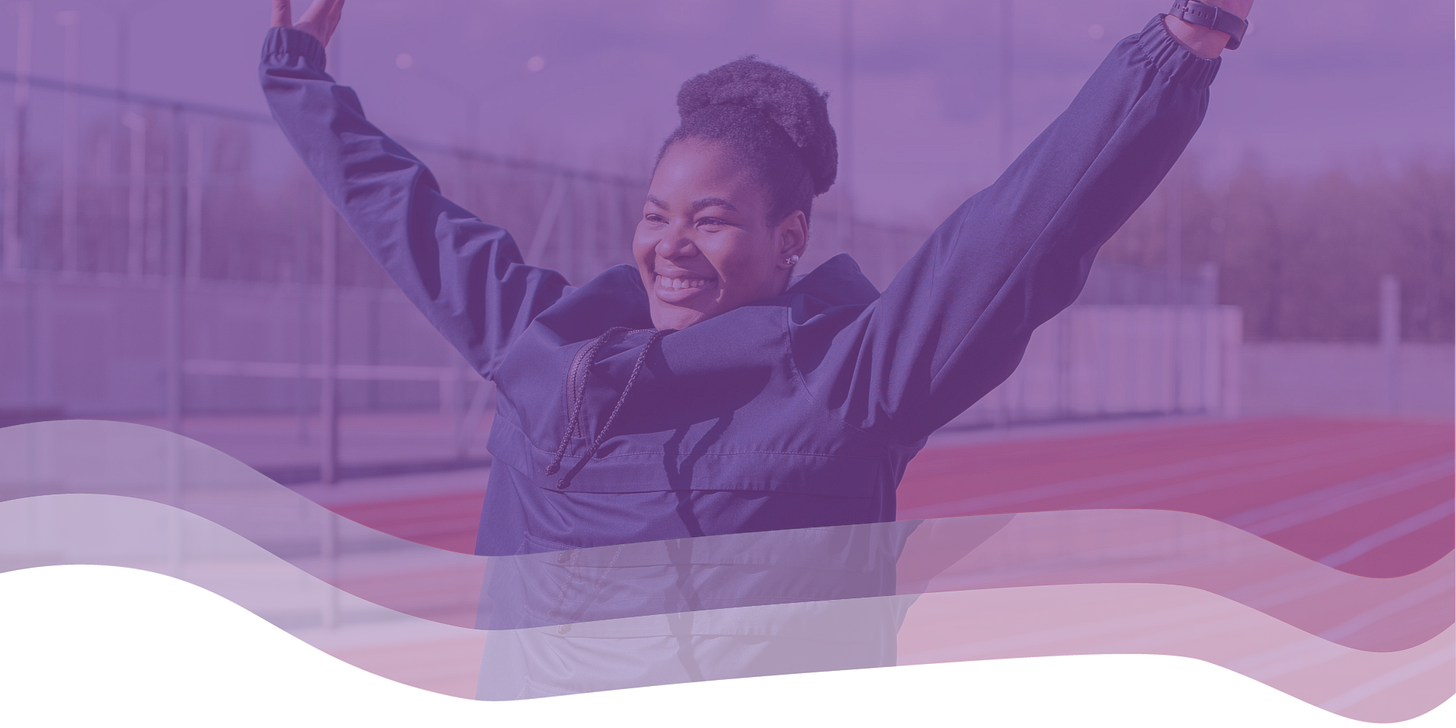 A woman raises her hands in celebrating, smiling, against the background of a running track