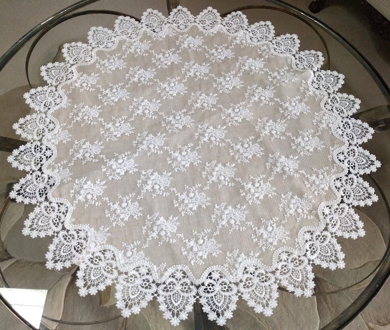 May include: A white round lace tablecloth with a delicate floral pattern. The tablecloth has a scalloped edge and is made of a sheer fabric.