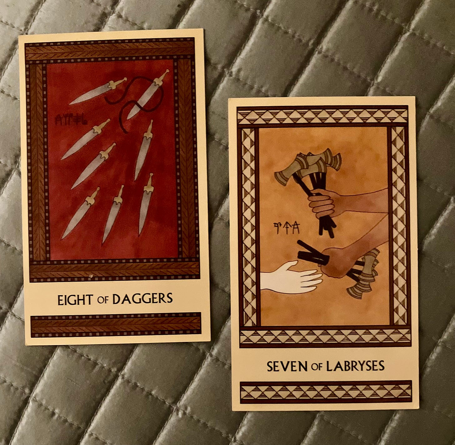 Two Minoan Tarot cards side by side on a silvery quilted fabric surface. The Eight of Daggers is painted in shades of dark red and brown. It shows eight daggers flying away from a cord that had held them tight but that has now been untied. The Seven of Labryses is painted in shades of deep yellow, cream, and tan. It shows a pair of hands holding seven labryses and handing some of them to a third hand.