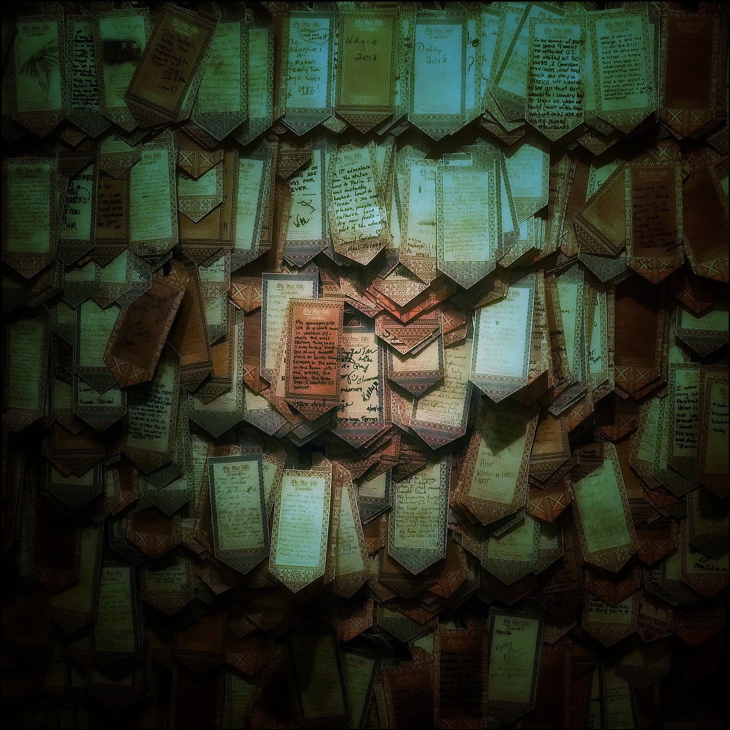 A murky photo of a wall to which hundreds of assorted tags have been affixed. The tags are in disorderly rows, overlapping one another, and each tag seems to include a note: a heading at the top and smaller text below, although neither the headline nor the text is legible.