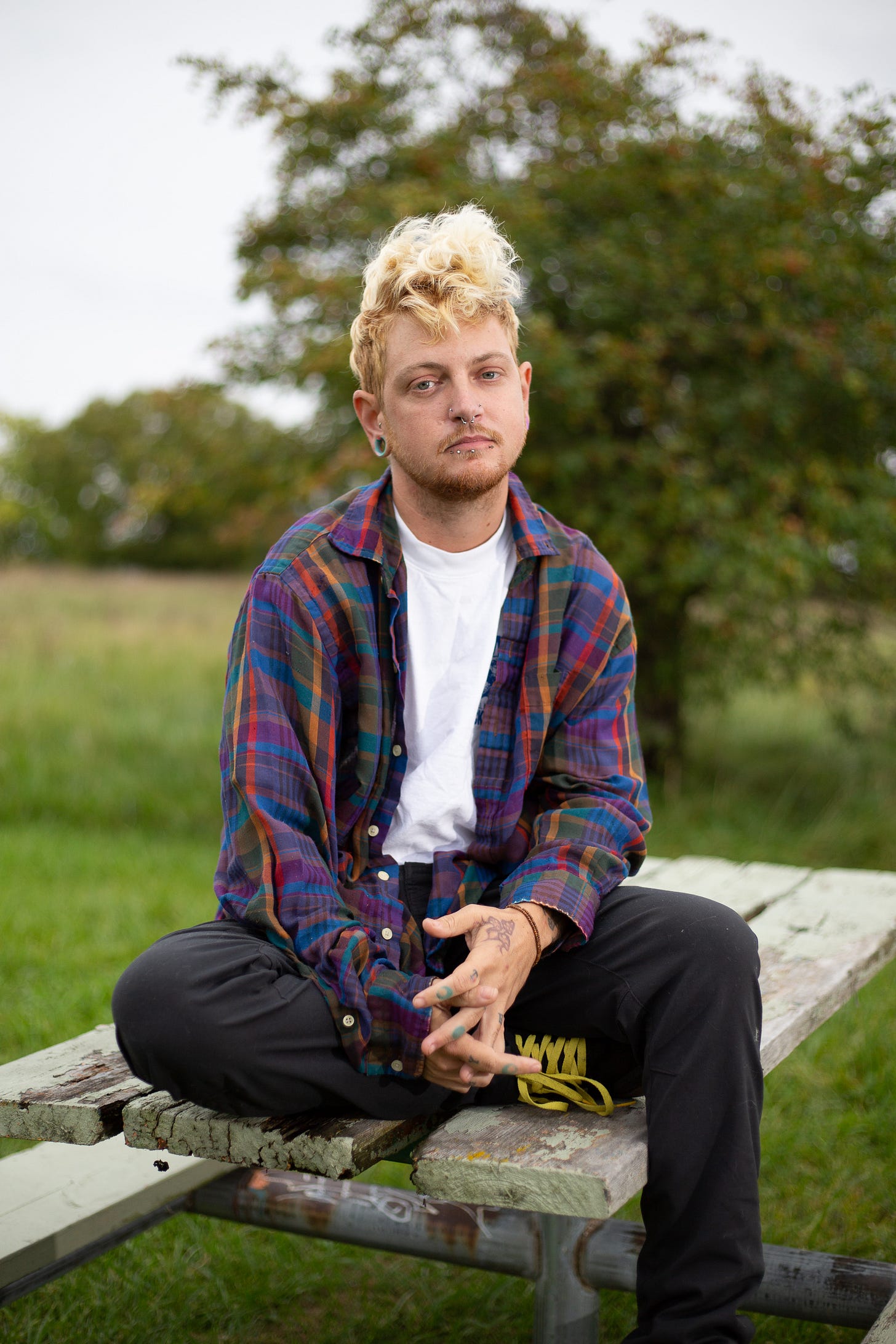Kaje sits on a park table outdoors. They are wearing black pants and a multi-colored plaid shirt. They have short blonde curly hair and tattoos on their hands.